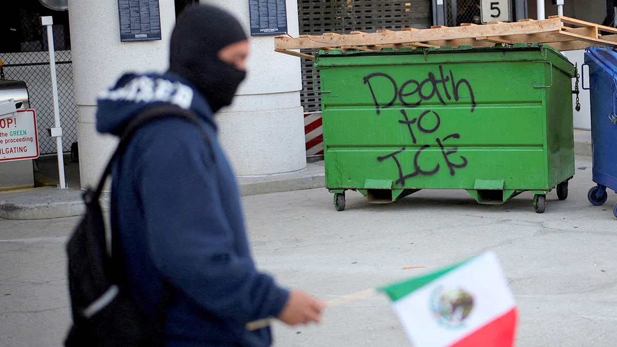 Person walks past while holding a Mexican flag by passing a dumpster that is spray painted